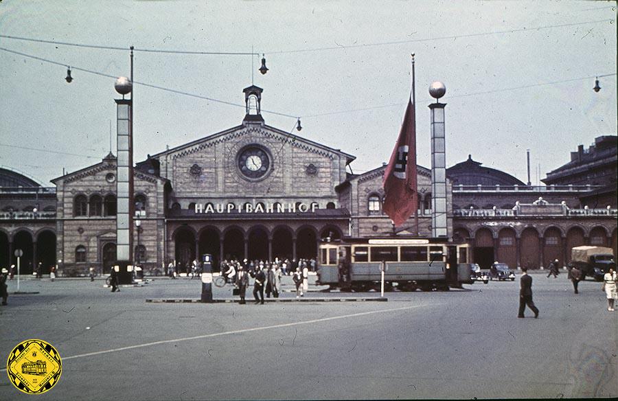 Das letzte Bild des intakten Bahnhofsgebäudes, dafür das erste Farbfoto von vor dem 2. Weltkrieg. Die großen Flaggen und martialischen Zusatzsäulen verheißen nichts Gutes.