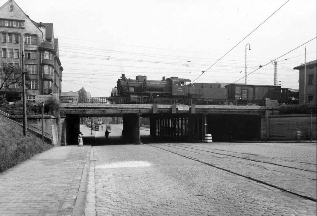 Die Dampflok BR 54 1655 auf der Lindwurmunterführung und im Hintergrund kommt ein A-Wagen: Betrieb auf der Lindwurmstraße im Jahr 1930. Auf der Bahnstrecke startet am 12. April 1927 der elektrische Betrieb.