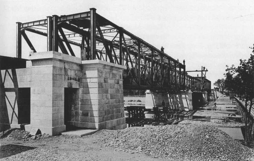 Der hölzerne Werkssteg (rechts im Bild noch zu sehen) wurde nach Fertigstellung der Brücke 1871 weiter flussabwärts, am Standort der heutigen Wittelsbacherbrücke, als provisorische Isarüberquerung neu errichtet.