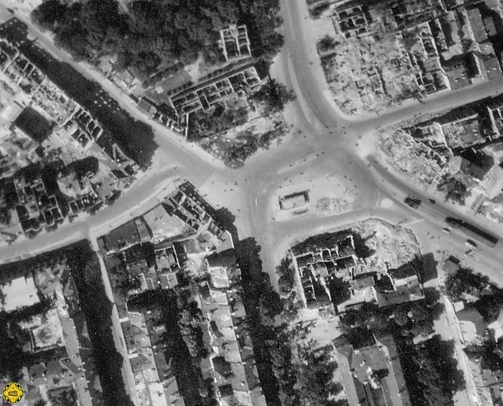 Luftaufnahme vom Rotkreuzplatz am 8. Juni 1945. Das Trambahnstationshaus (in der Bildmitte) hat kein Dach mehr. Der gesamte Platz ist am 7.1.1945 zerstört worden. Detail am rechten Rand: dort erkennt man zwei Dampflokomotiven und einen Reichsbahn-Steuerwagen der von der Reichsbahn betriebenen Hilfsbahn Linie IV vom Stiglmaierplatz kommend, die hier am Anwesen 158 der Nymphenburgerstraße ihr Kohlelager hatten.