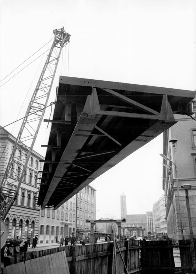 1966 11 19 Aufbau Stahlhochbrücke Von Der Tann Str Ludwigstr (16) Die ersten Brückenelemente für den Mittelteil sind 30m lang und 55 Tonnen schwer. Danach werden mit einem zweiten Autokran die weiteren Brückenelemente in beide Richtungen bis zu den Rampen eingesetzt. Am Abend des 19.November 1966 sind dies Arbeiten komplett abgeschlossen. 